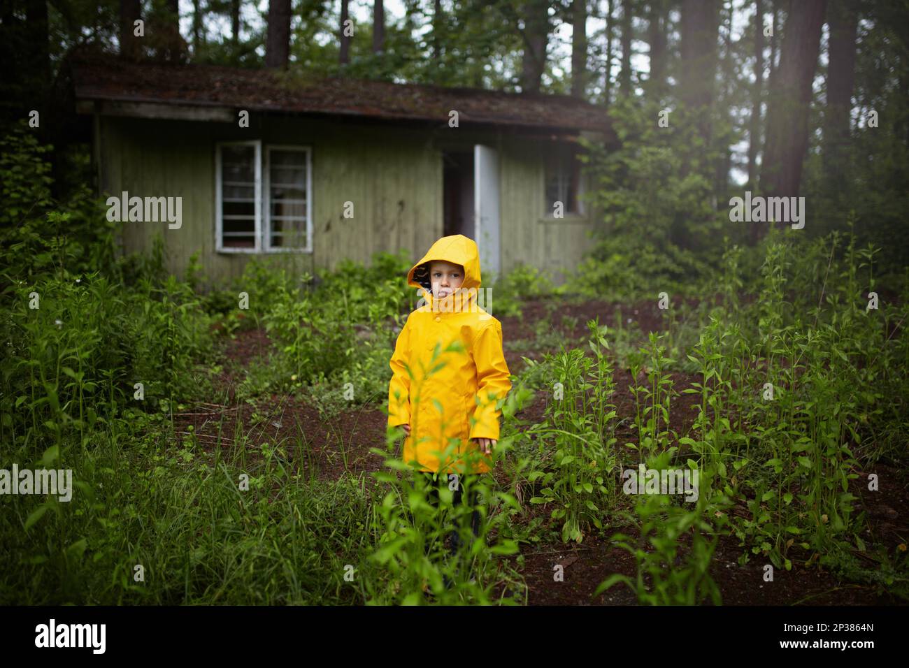 Ein Kind in einem gelben Mantel, das im Wald vor einem alten Holzhaus steht. Ein 6-jähriger Junge Stockfoto