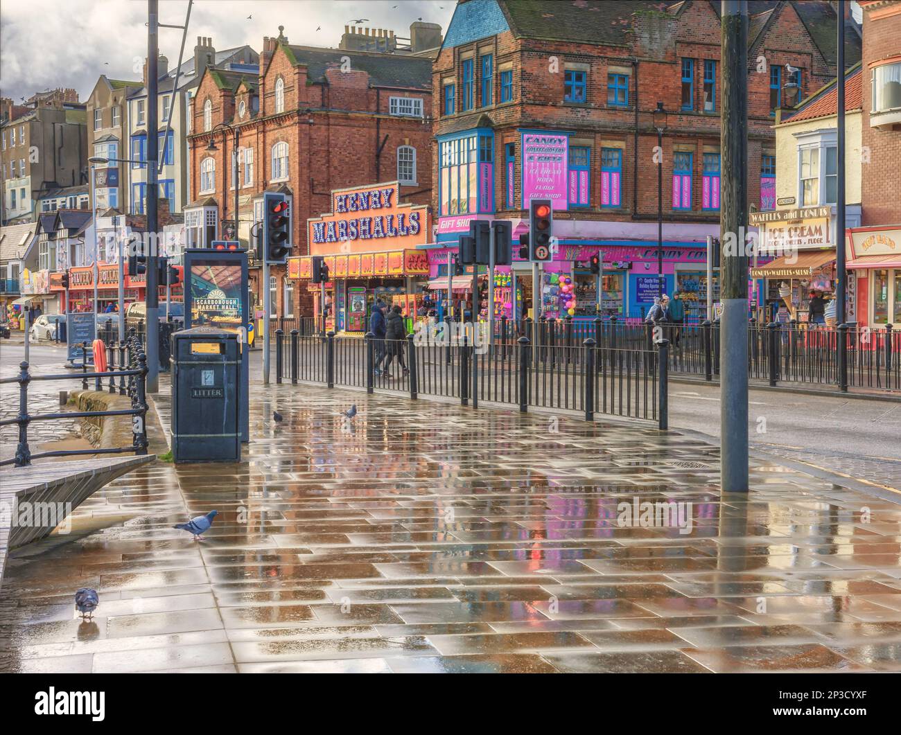 Eine Regendusche hinterlässt einen nassen Bürgersteig mit Reflexionen. Tauben stehen im Vordergrund und die Straße ist von Geschäften und Vergnügungsparks gesäumt. Stockfoto