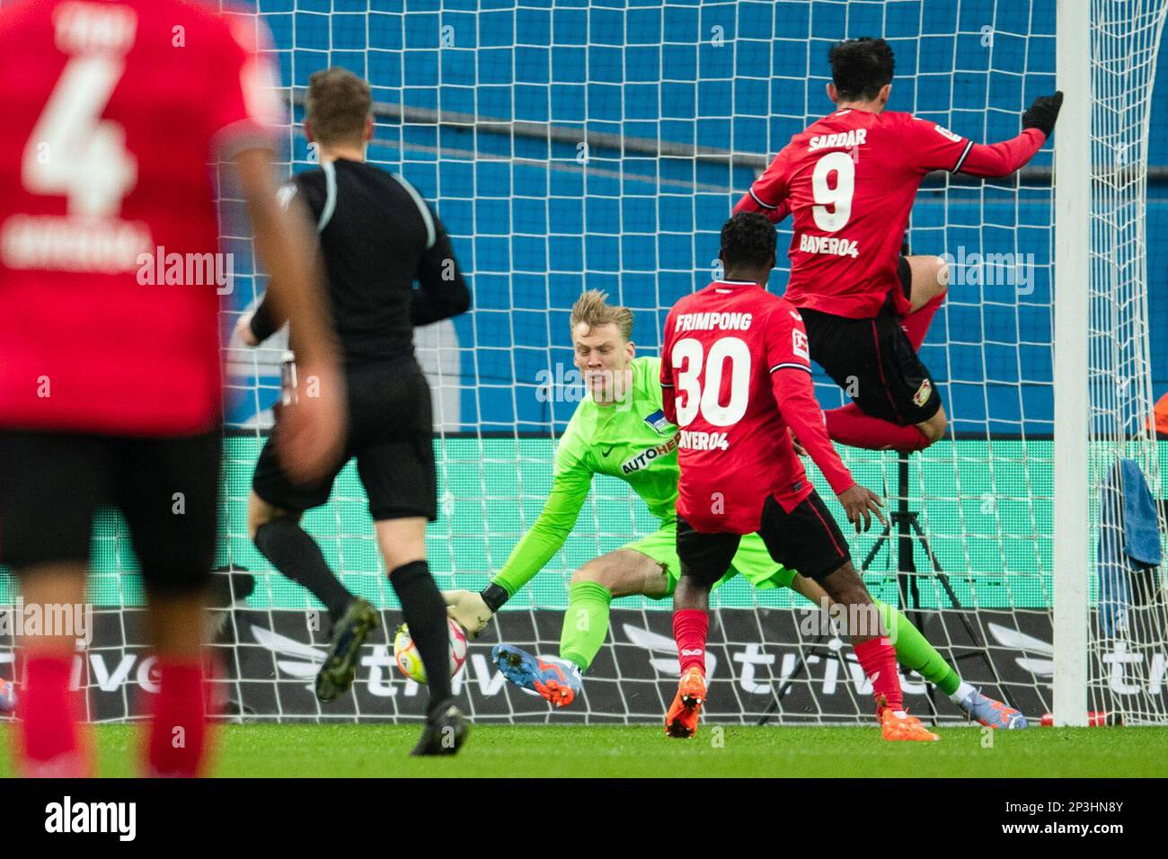 05. März 2023, Nordrhein-Westfalen, Leverkusen: Fußball: Bundesliga, Bayer Leverkusen - Hertha BSC, Spieltag 23, BayArena. Leverkusens Jeremie Frimpong (2. von rechts) trifft gegen Hertha Torhüter Oliver Christensen (M) und schafft es 2:0. Foto: Marius Becker/dpa - WICHTIGER HINWEIS: Gemäß den Anforderungen der DFL Deutsche Fußball Liga und des DFB Deutscher Fußball-Bund ist es verboten, im Stadion aufgenommene Fotografien und/oder das Spiel in Form von Sequenzbildern und/oder videoähnlichen Fotoserien zu verwenden oder verwenden zu lassen. Stockfoto