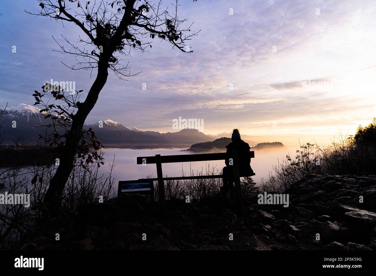 2022 12 31, Bled, Slowenien: Die Frau, die auf einer Bank saß und auf den See blickte, blutete während des Sonnenaufgangs, vollständig von Nebel bedeckt Stockfoto