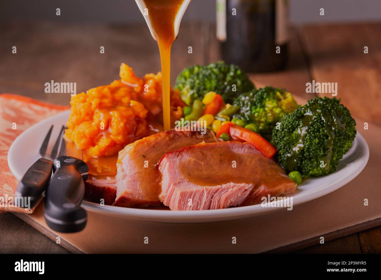 Gammon-Dinner mit Gemüse, Süßkartoffeln und dicker Soße, die darüber gegossen wird. Stockfoto