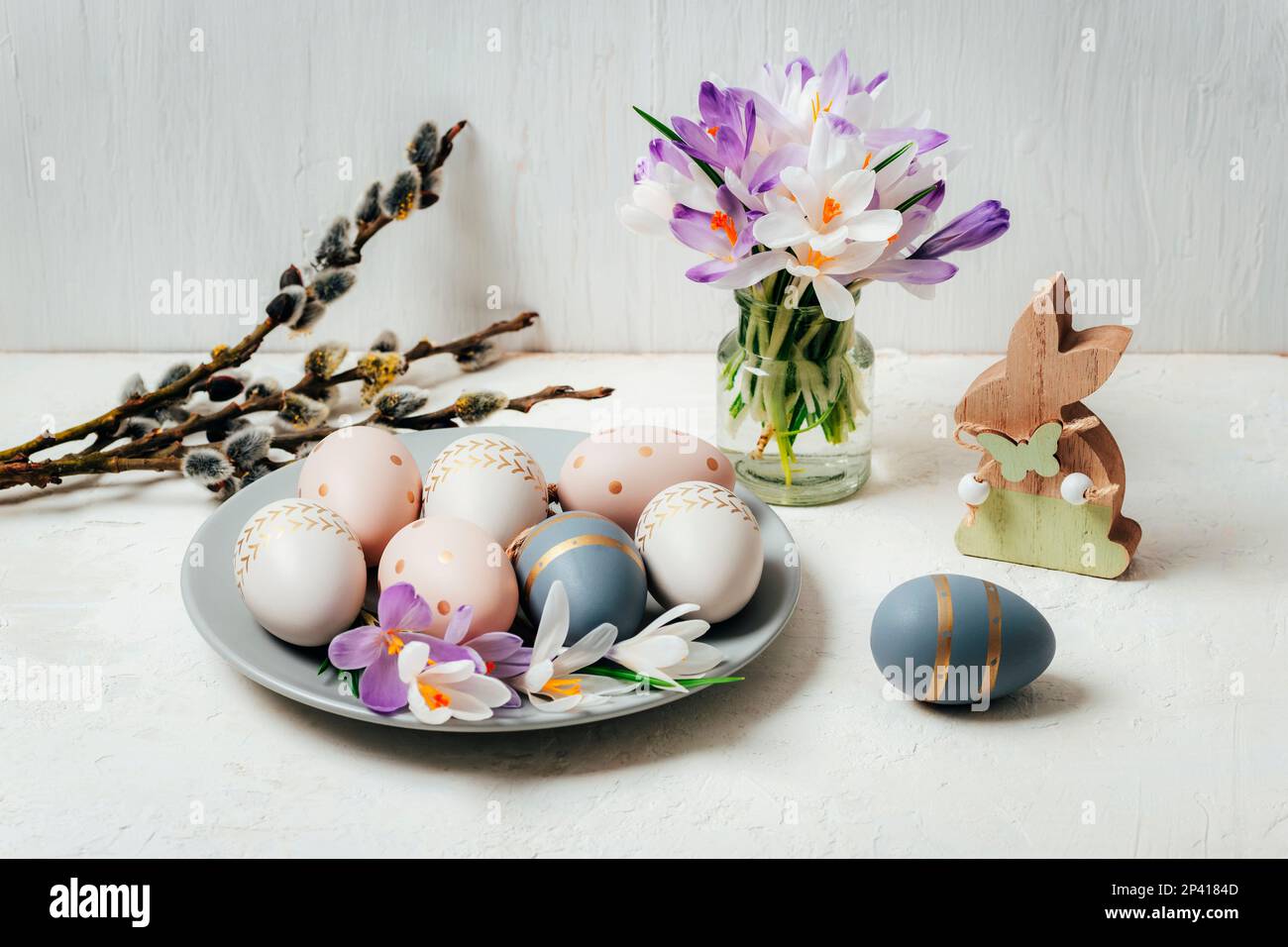 Grauer Teller mit bemalten Ostereiern und Holzhasen, Krokusblumen auf weißem Tisch. Ostergrußkarte. Stockfoto