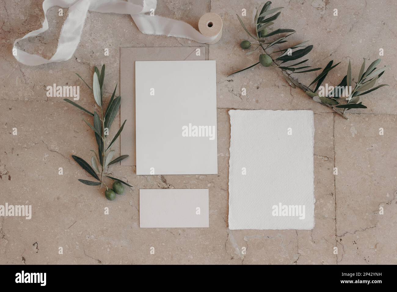 Italienischer Sommer Hochzeit Schreibtischset. Mockup-Szene mit leerem Grußpapier, RSVP-Karten auf beigefarbenem Marmorfliesen-Hintergrund. Grüne Olivenbaumzweige Stockfoto