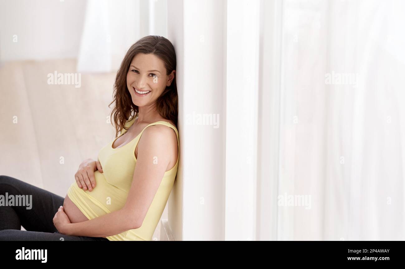 Ich bin gesund, glücklich und schwanger. Eine junge schwangere Frau, die auf ihrem Wohnzimmerboden sitzt. Stockfoto