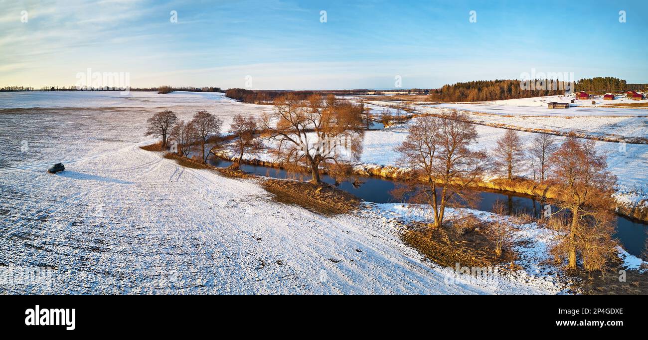 Der Fluss fließt in verschneiten Winterfeldern in der Wildnis bei Sonnenuntergang. An kalten Abenden schlängeln sich die eisigen Landschaften. Blick auf Bäume am Flussufer aus der Vogelperspektive. Stockfoto