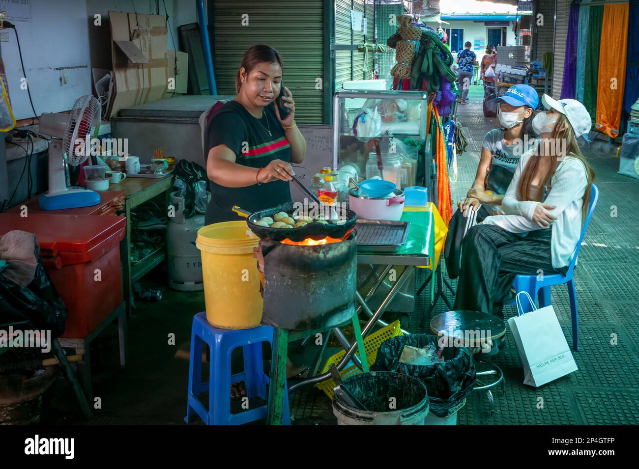 Eine Straßenverkäuferin auf dem Central Market in Phnom Penh, Kambodscha, chattet auf ihrem Handy, während sie süße Kuchen frittiert und zwei junge Kunden warten. Stockfoto
