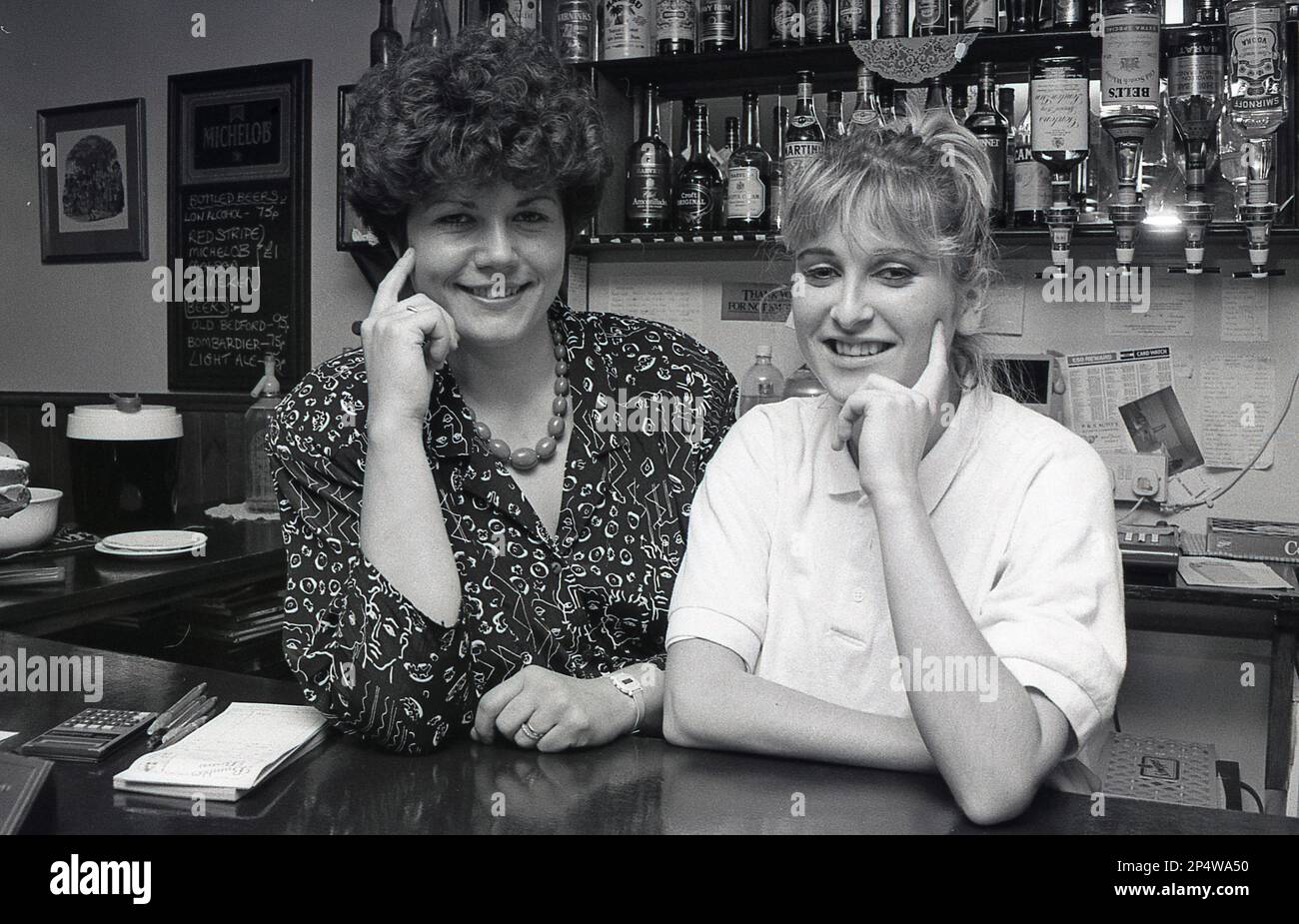 1980er, zwei junge Barkeeper posierten für ihr Foto hinter der Bar im Bistro oder in der Café-Bar ... links auf dem Bild ein kleines Kreidetafel, auf dem der Preis für Bier aufgeführt ist ... unter £1,00 pro Person, oh, das waren die Tage! Stockfoto