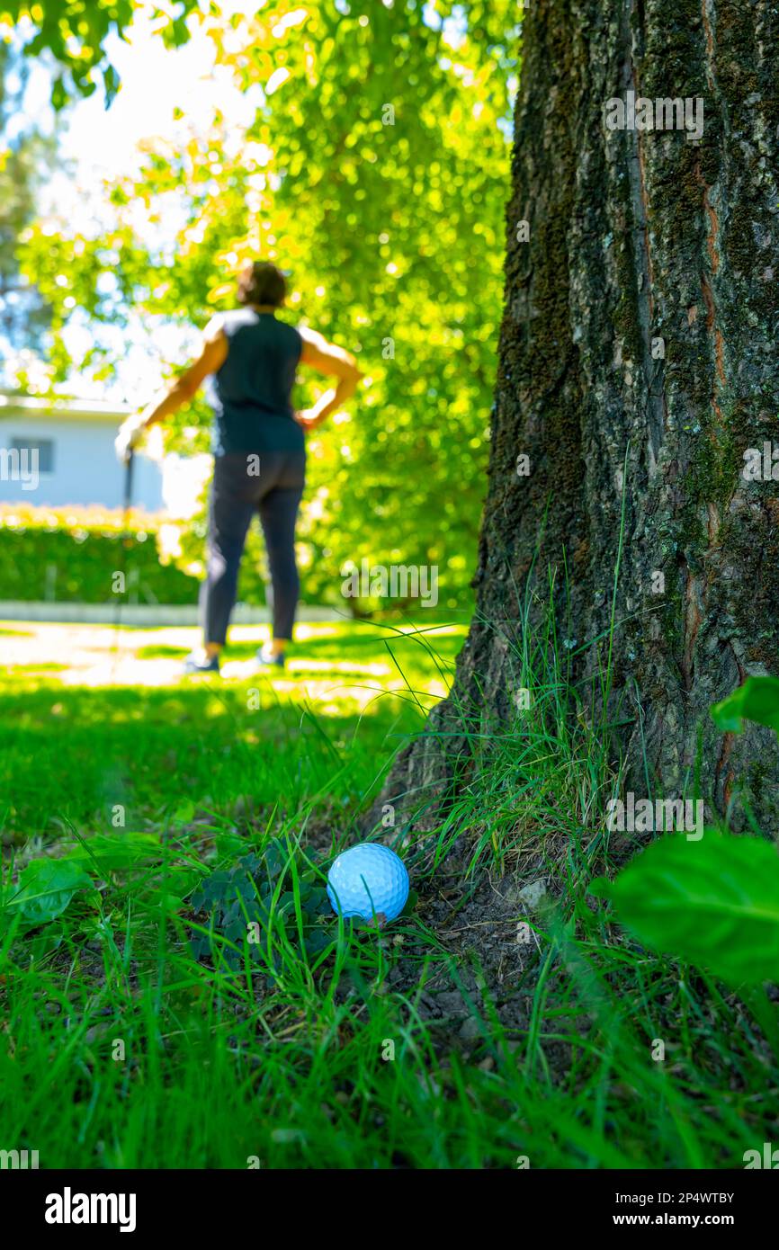Golfball auf dem rauen Gras und Golfer mit Golfclub auf der Suche nach dem Golfball in den Bäumen an einem sonnigen Tag in der Schweiz. Stockfoto