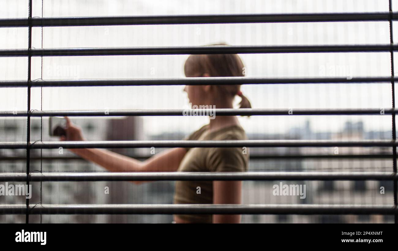 Influencer Teenager Mädchen in der Stadt hinter offenen horizontalen Aluminium Metall schwarzen Jalousien, mit einem Smartphone. Nahaufnahme, Fensterhintergrund. Rückansicht, br Stockfoto