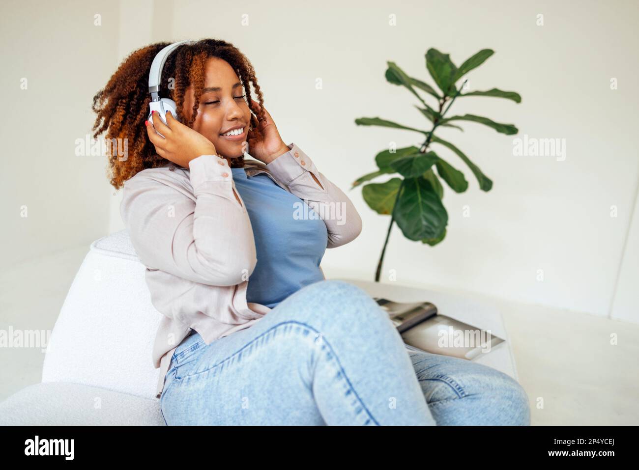 Körperpositive, kurvige afro-amerikanische Frau in Kopfhörern auf einem hellen Sofa zu Hause. Eine dunkle Frau in legerer Kleidung schloss die Augen und genießt es Stockfoto