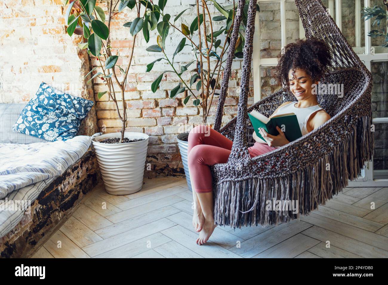 Ein junges, wunderschönes afro-amerikanisches Mädchen liest in einem Korbstuhl. Dunkelhäutiger Teenager in legerer Kleidung ruht zu Hause aus. Weibliches Teenager-Lächeln Stockfoto