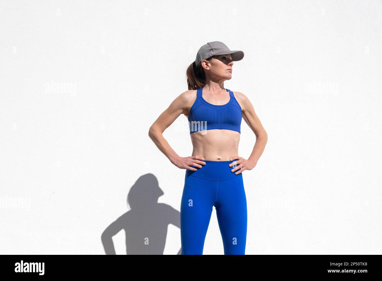Fit sportliche Frau, die mit den Händen auf der Hüfte steht, eine Baseballmütze trägt und draußen in der Sonne trainiert Stockfoto