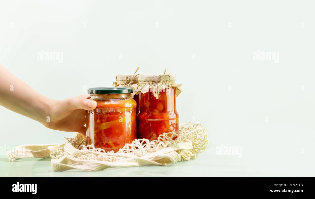 Probiotische Lebensmittel. Nachhaltiges Stillleben mit eingelegtem oder fermentiertem Gemüse mit der Hand einer Frau. Lecho und Ajvar in Glasgefäßen in einem Netzbeutel auf blauem Backgr Stockfoto