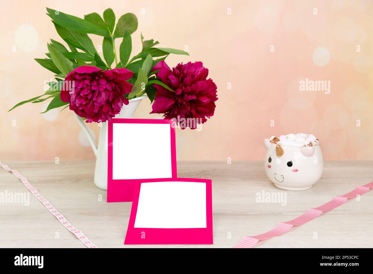 Modell für quadratische Bereiche mit rosafarbenem Rahmen. Pfingstrosen (Paeonia) in einer weißen Zinnvase. Ein Einhorn-Becher mit Marshmallows. Heller Hintergrund in Pfirsich und Pink bis Stockfoto