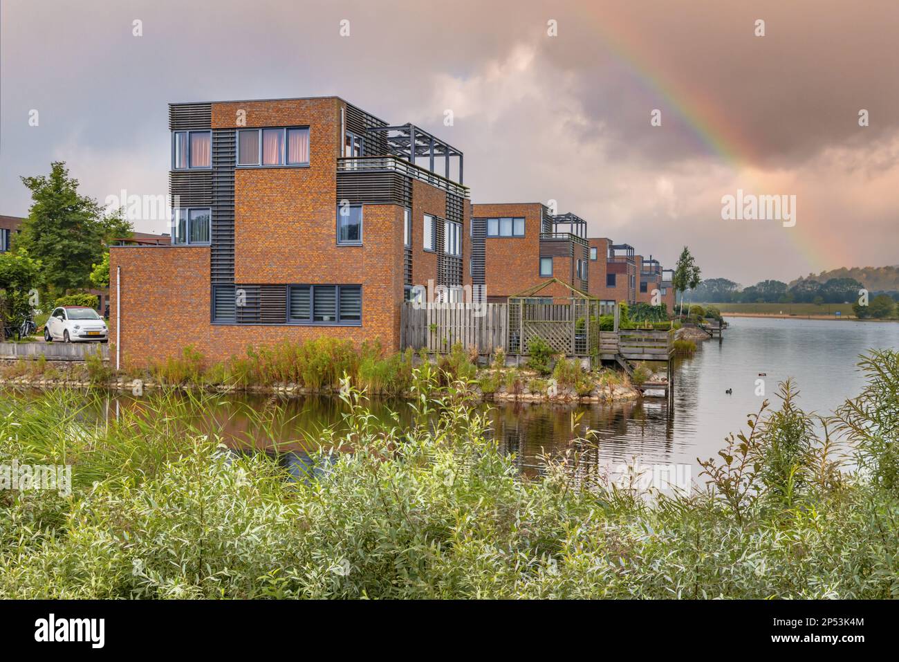 Häuser am Wasserrand in Wohngebieten in den Niederlanden. Unter bewölktem Himmel mit Regenbogen. Heerhugowaard, Niederlande. Stockfoto