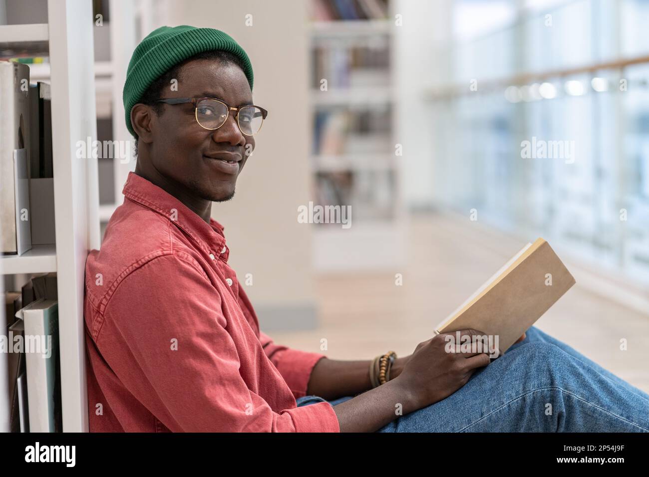 Ein schwarzer Student, der auf dem Boden neben einem Bücherregal sitzt und ein Buch in der Hand hält, schaut in die Kamera Stockfoto