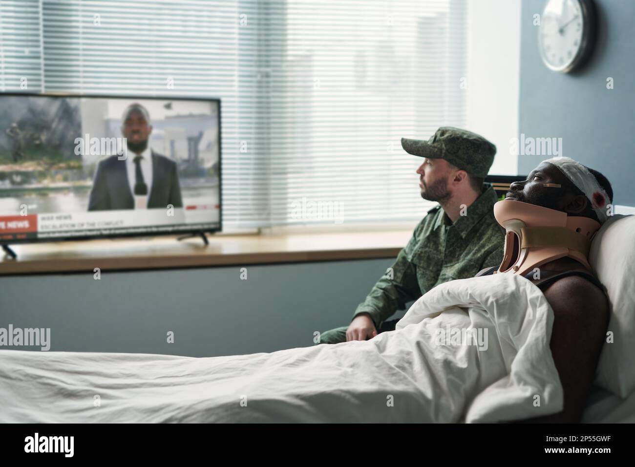 Seitenansicht des verletzten Mannes und seines Freundes in Militäruniform, der Nachrichten im fernsehen ansieht, während er vor dem Bildschirm in der Krankenhausabteilung sitzt Stockfoto