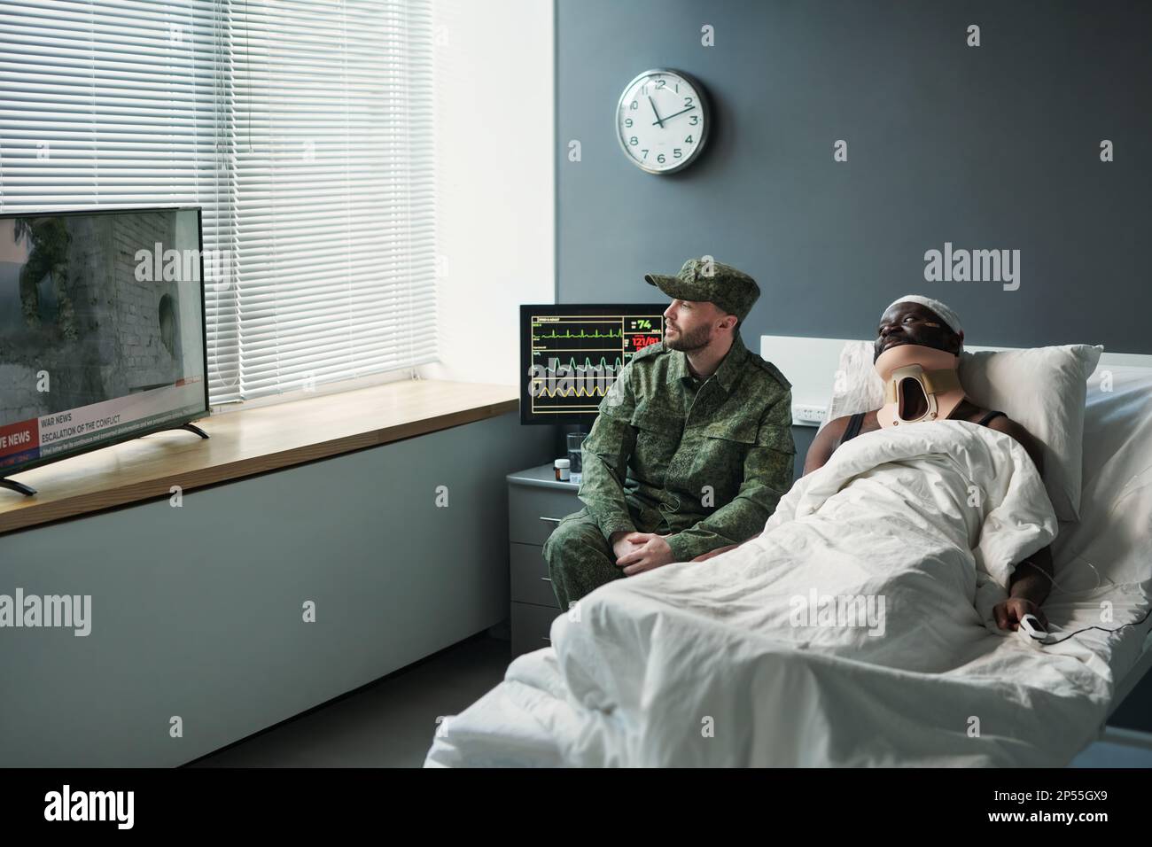 Junger Soldat in Militäruniform sitzt am Bett eines verletzten Freundes mit Verbandskopf, während beide im Krankenhaus die neuesten Nachrichten im fernsehen sehen Stockfoto