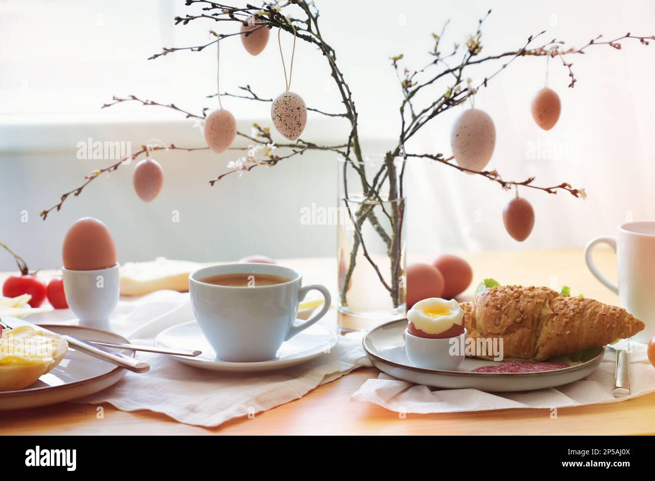Osterfrühstück in der Morgensonne mit Kaffee und Croissant auf einem Tisch am Fenster, Dekoration aus natürlichen Eiern, die an den Ästen hängen, Kopie Stockfoto