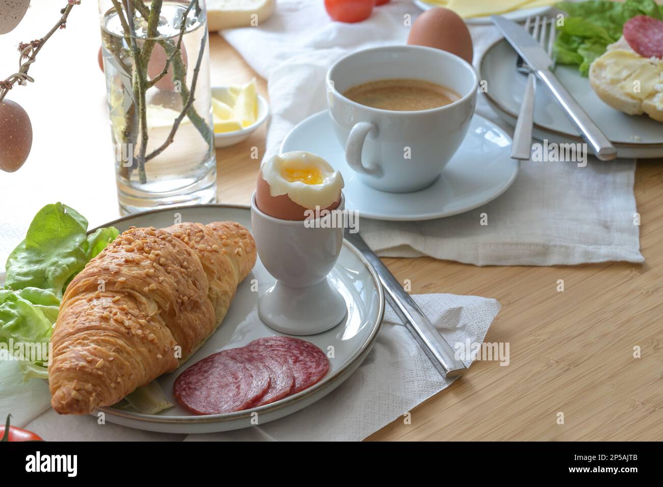 Frühstück oder Brunch mit Croissant, gekochtem Ei, Wurst, Käse und Kaffee auf leichten Servietten und einem Holztisch, ausgewählter Fokus, schmale Feldtiefe Stockfoto