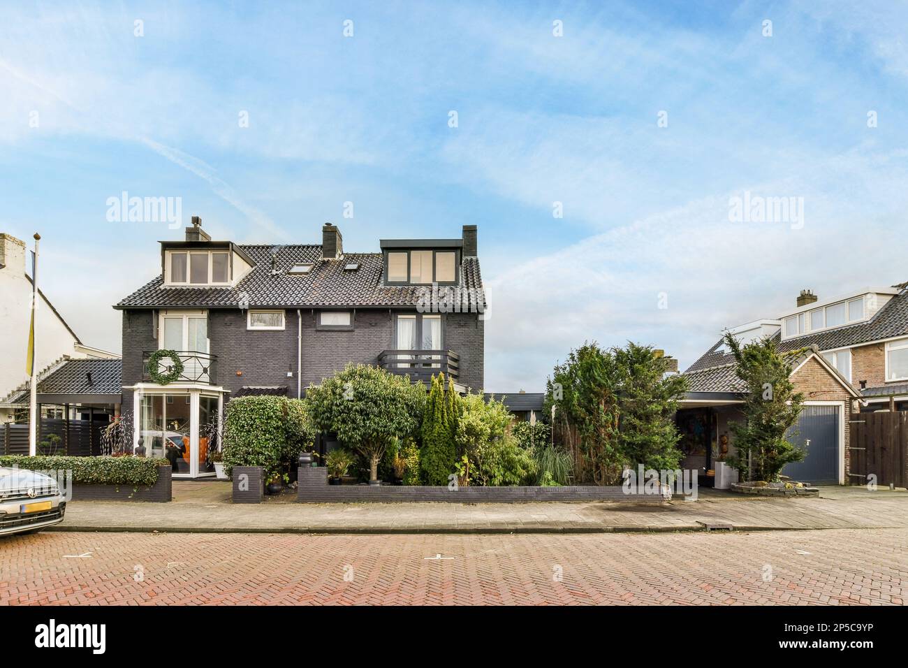 Ein Haus mit einem Auto in der Einfahrt daneben auf einer gepflasterten Straße mit Bäumen und Büschen Stockfoto