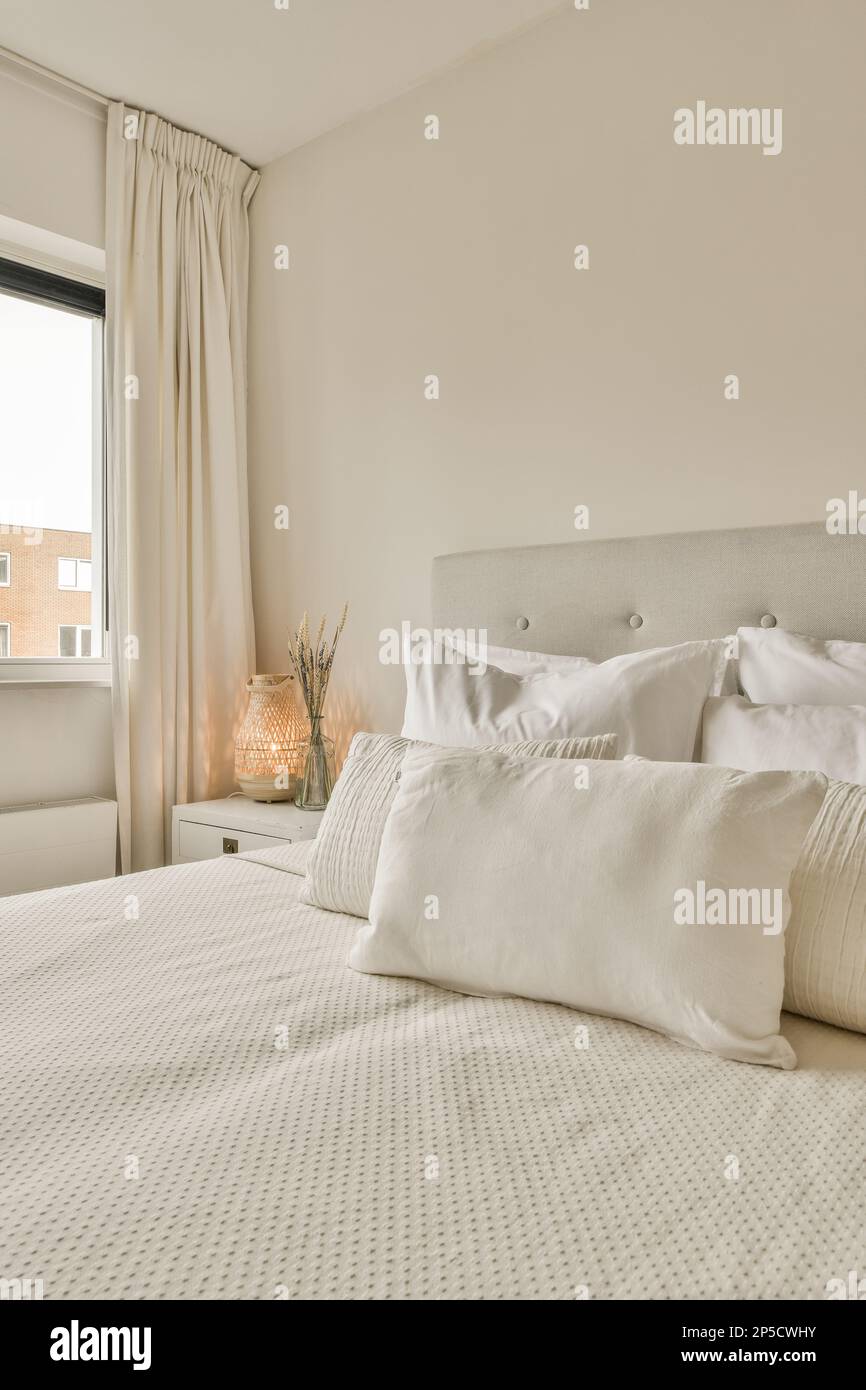 Ein Bett mit weißen Laken und Kissen in einem kleinen Zimmer neben einem Fenster, das von der Sonne beleuchtet wird Stockfoto