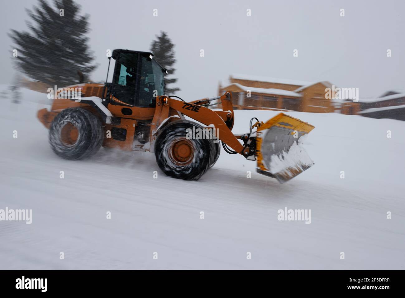 Während eines Schneesturms in Mammoth Lakes, Kalifornien, fährt ein gelber Traktor mit Schneeketten an einem im Bau befindlichen Haus vorbei. Stockfoto