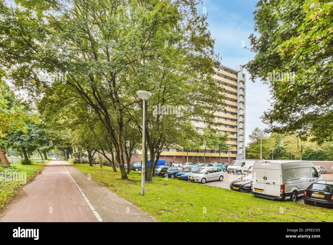 Amsterdam, Niederlande - 10. April 2021: Autos parken auf einer Straße vor einem Apartmenthaus mit grünen Bäumen und blauem Himmel Stockfoto