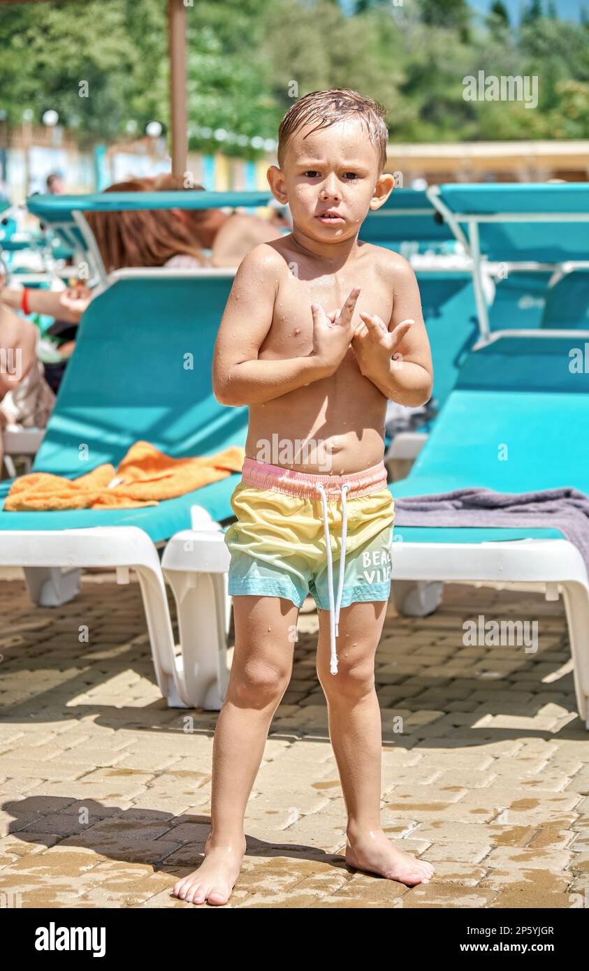 Vorschulkind mit nassen Haaren weht nach dem Schwimmen im Pool vom Wind. Der Junge steht an sonnigen Tagen vor Kälte vor Liegestühlen im Wasserpark Stockfoto