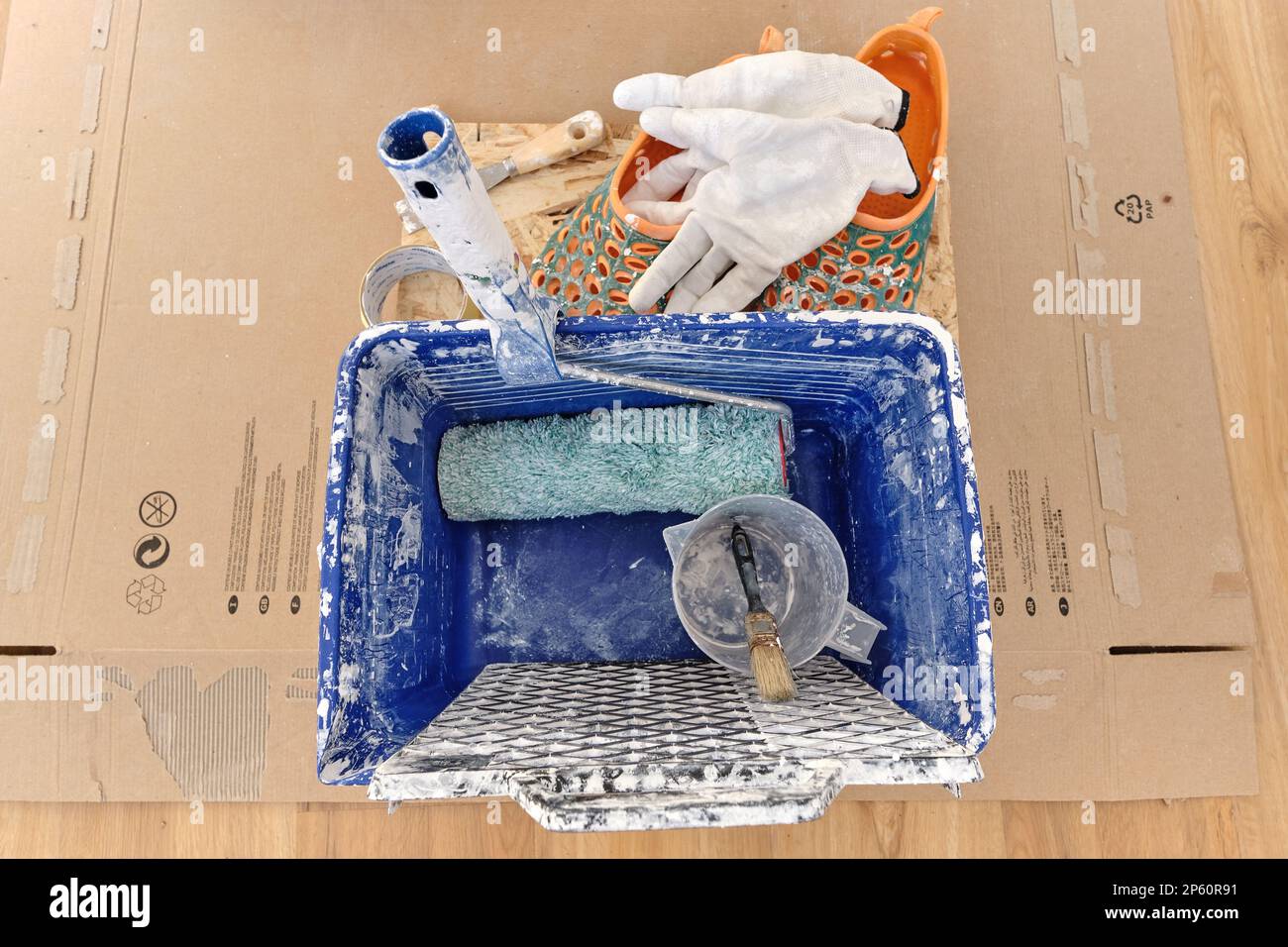 Blauer quadratischer Kunststoffeimer, der auf Pappe steht, von weißer, trockener Farbe verschmutzt, einschließlich Basiswerkzeug wie Raster, Rolle, Messbecher, Pinsel Stockfoto
