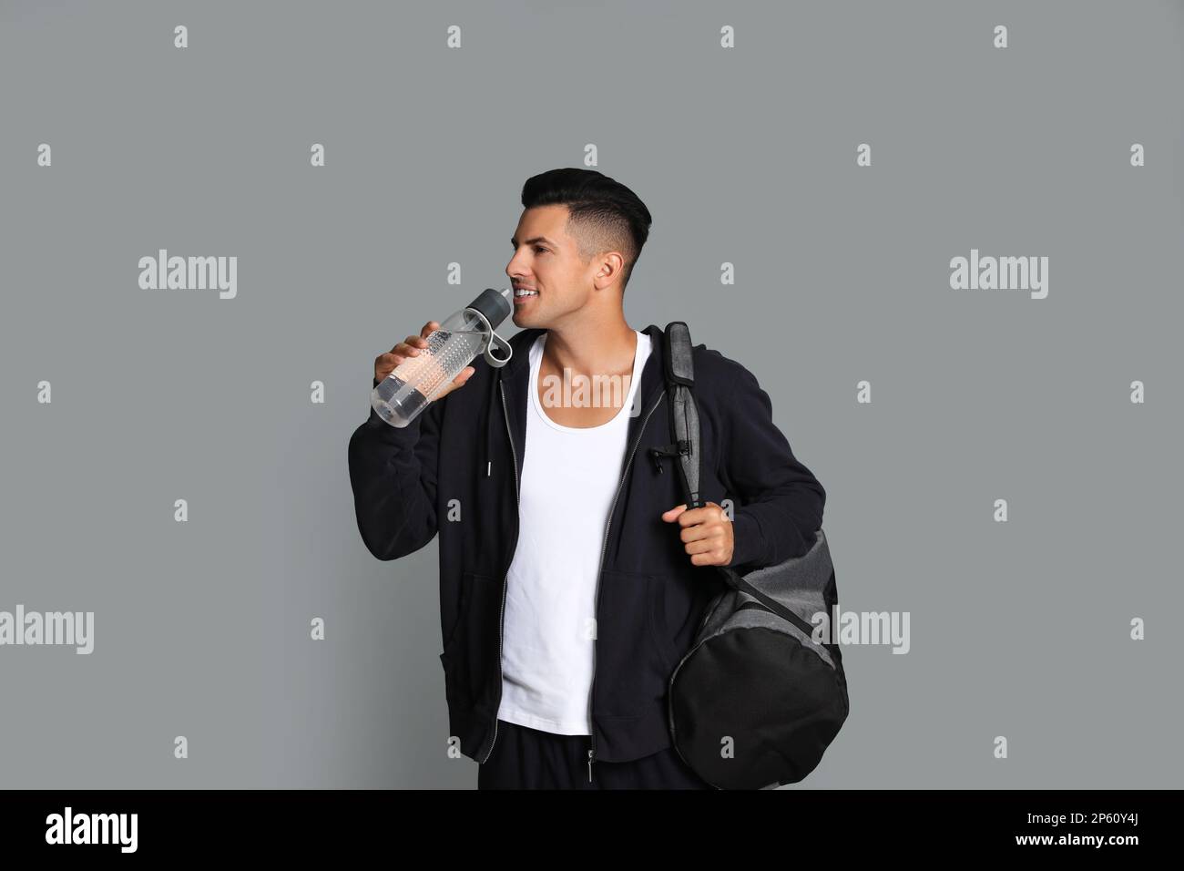 Hübscher Mann mit Sporttasche, der Wasser auf grauem Hintergrund trinkt Stockfoto