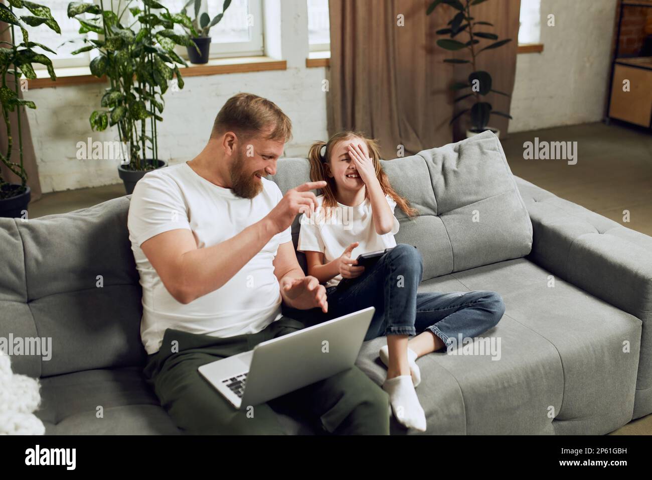 Glücklicher, lächelnder, reifer Mann, Vater, der auf der Couch mit einem Laptop sitzt, zu Hause aus der Ferne arbeitet und mit seiner kleinen Tochter spielt Stockfoto