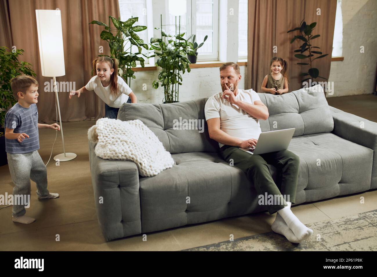 Ein erwachsener Mann, Vater, der zu Hause auf der Couch sitzt und per Fernzugriff an einem Laptop arbeitet. Kleine Kinder, die lautstark spielen und ihn belästigen Stockfoto