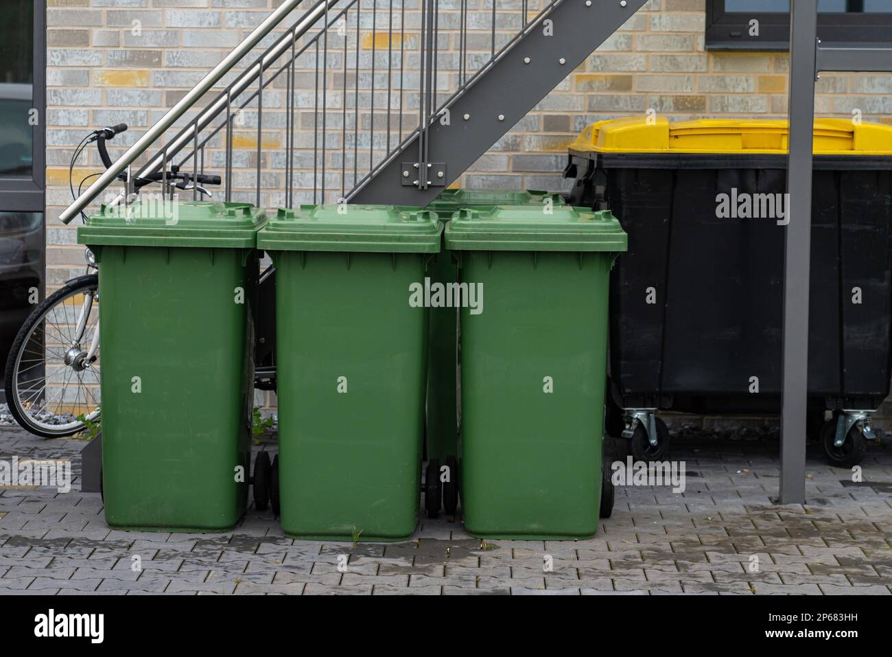 Drei grüne Mülltonnen vor einem Wohngebäude. Fahrrad und schwarzer Abfalleimer unter der Metalltreppe. Stockfoto