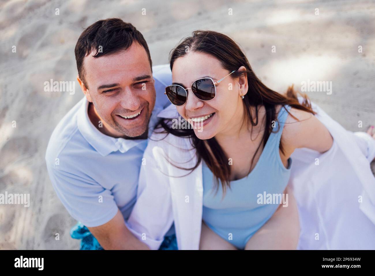 Ein weißes Paar, das sich an einem Sandstrand liebt. Ein junger Mann sieht eine schöne Frau mit Sonnenbrille liebevoll an. Freundin und Freund lächeln sich an. Stockfoto