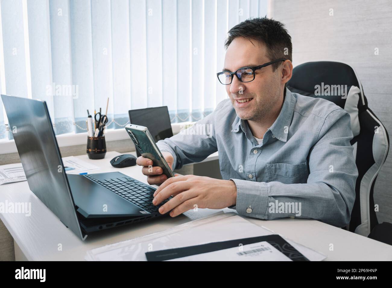Moderne Technik, Lifestyle, gute Nachrichten. Ein Mann sitzt am Arbeitsplatz mit einem Laptop und nutzt ein Smartphone lächelt, liest angenehme sms, bekommt ein Angebot, chattet online Stockfoto