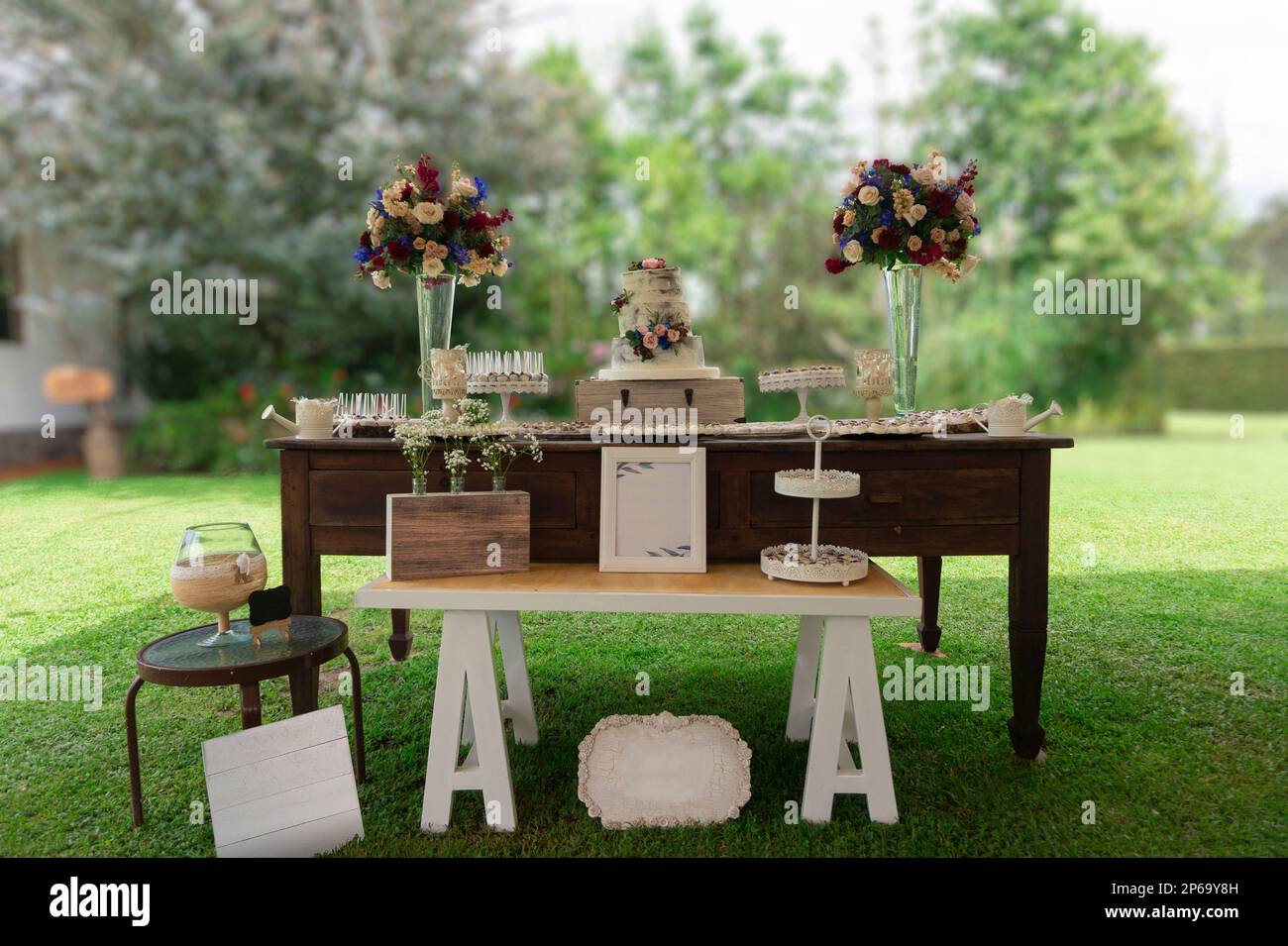 Hochzeitstorte auf einem alten Holztisch in einem Garten mit grünen Bäumen an einem sonnigen Tag, dekoriert mit Rosensträußen und Tellern Stockfoto