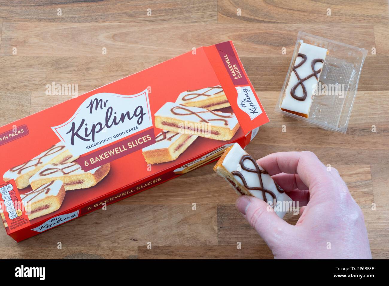 Die Hand eines Mannes, der ein Mr Kipling Bakewell Slice hält, mit einer Packung Scheiben im Hintergrund. UK. Konzept: Snacks, ungesunde Ernährung Stockfoto