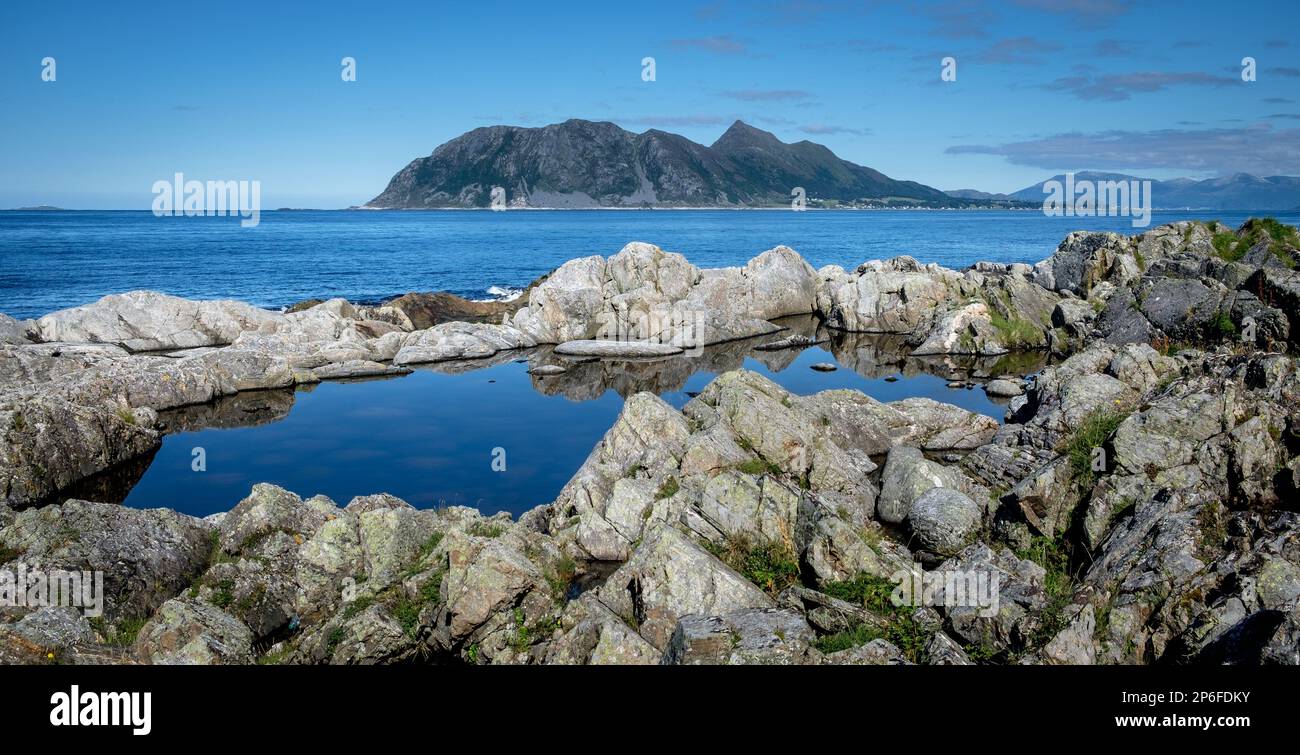 Eine ruhige Küstenlandschaft mit klarem, blauem Wasser, das Felsformationen reflektiert, und einer fernen bergigen Insel unter blauem Himmel. Stockfoto