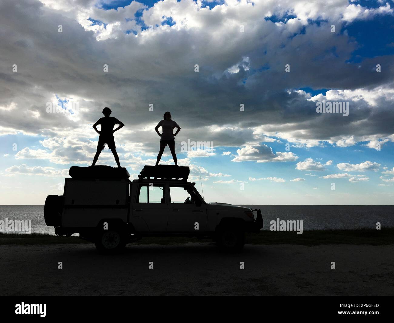 2 junge Erwachsene stehen auf einem Jeep als Silhouetten, die menschliche Figuren spielen. Sturmwolken am Himmel. Makgadikgadi Salzwannen, Botsuana, Afrika Stockfoto