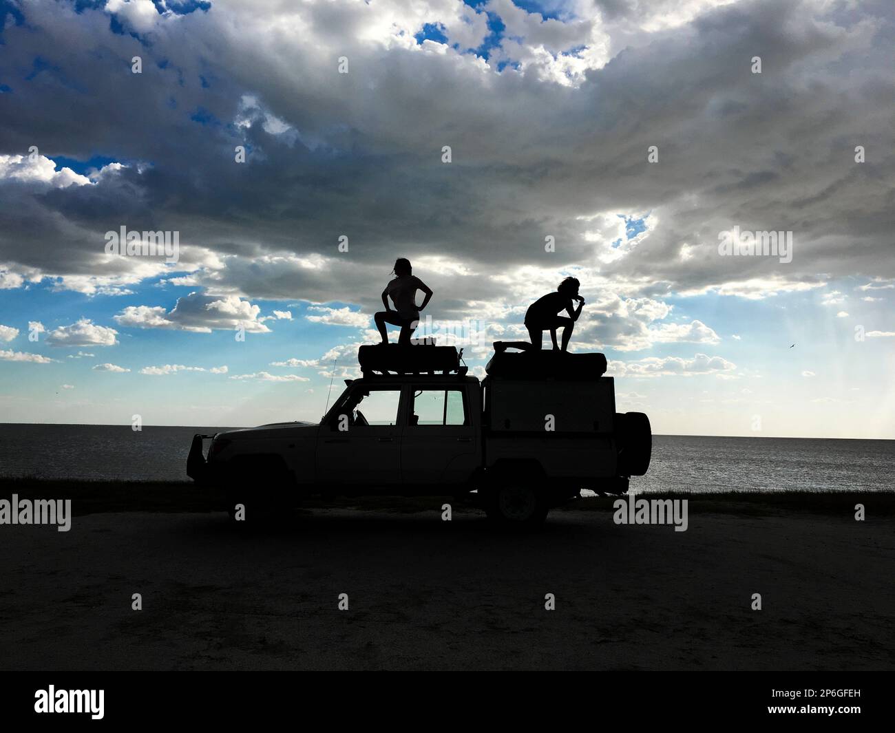 2 junge Erwachsene stehen auf einem Jeep als Silhouetten, die menschliche Figuren spielen. Sturmwolken am Himmel. Makgadikgadi Salzwannen, Botsuana, Afrika Stockfoto