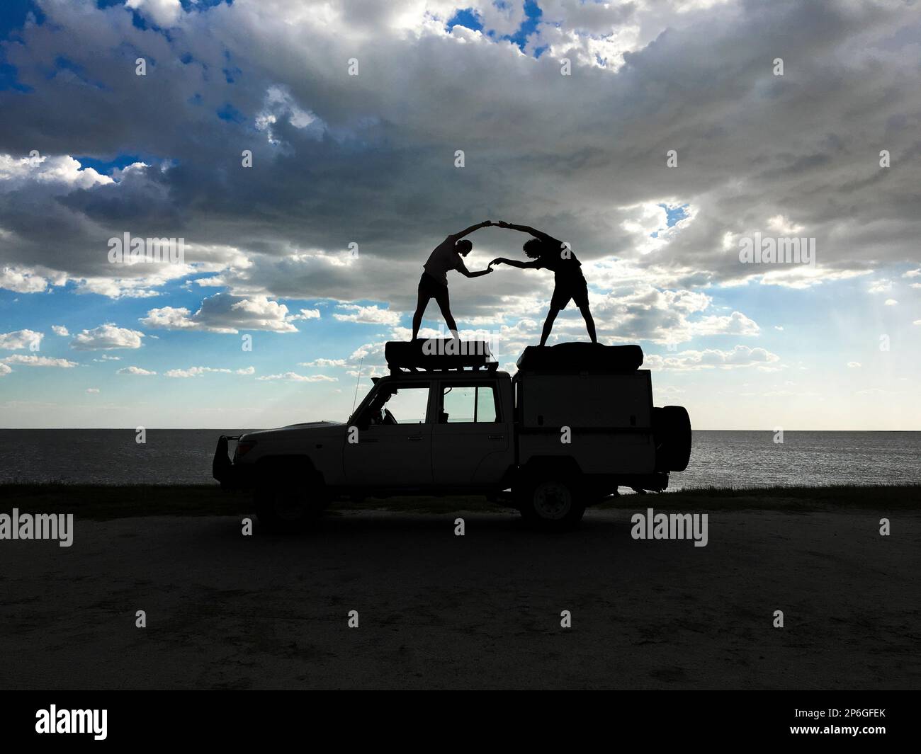 2 junge Erwachsene stehen auf einem Jeep als Silhouetten, die menschliche Figuren spielen. Sturmwolken am Himmel. Makgadikgadi Salzwannen, Botsuana, Afrika Stockfoto