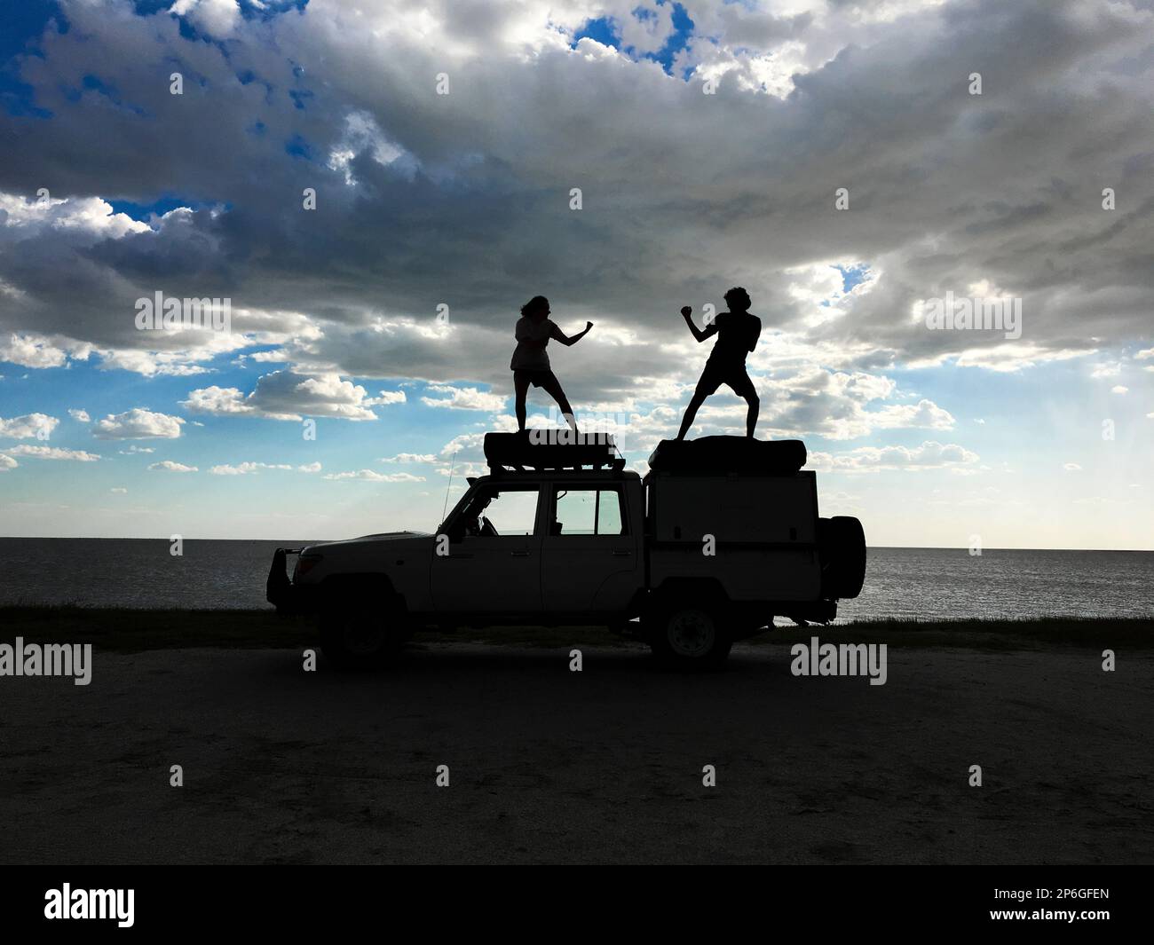 2 junge Erwachsene stehen auf einem Jeep als Silhouetten, die menschliche Figuren spielen. Sturmwolken am Himmel. Makgadikgadi Salzwannen, Botsuana, Afrika Stockfoto