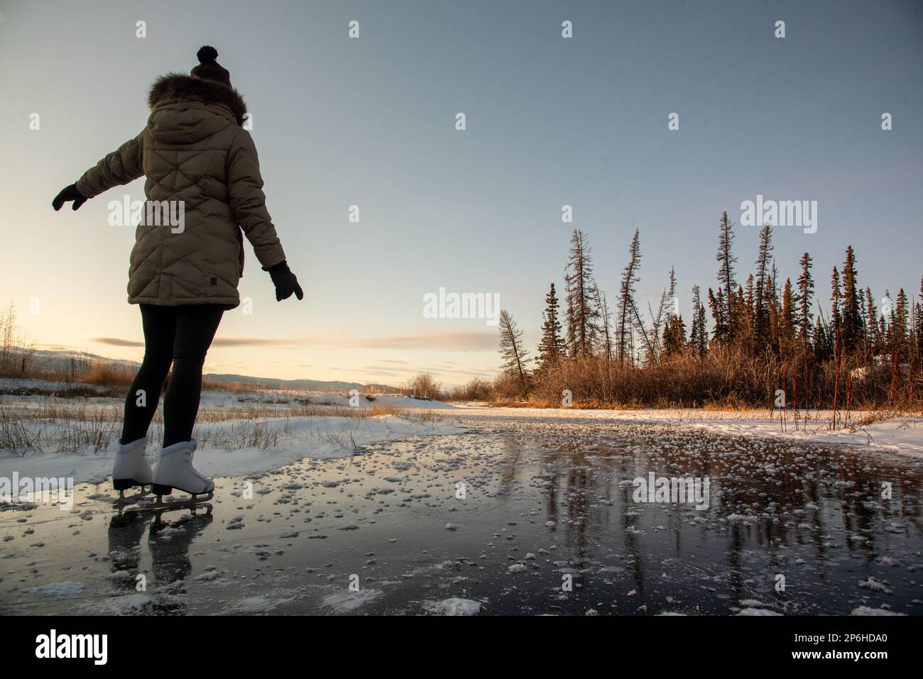 Frau, eine Person, die im Winter bei Sonnenuntergang auf einem gefrorenen See mit borealem Wald im Hintergrund skatert. Stockfoto