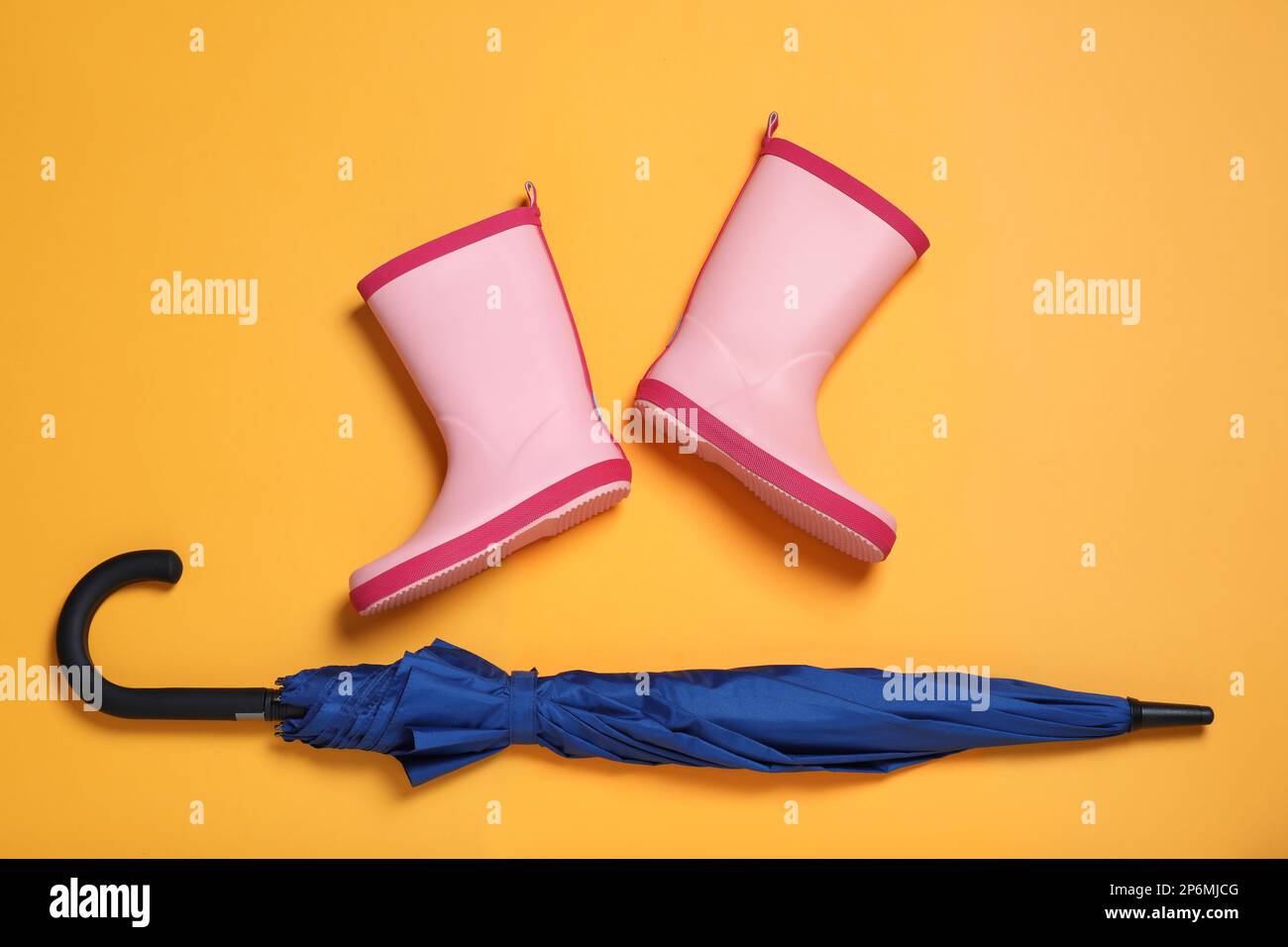 Pinkfarbene Gummistiefel neben blauem Regenschirm auf orangefarbenem Hintergrund, flach liegend Stockfoto