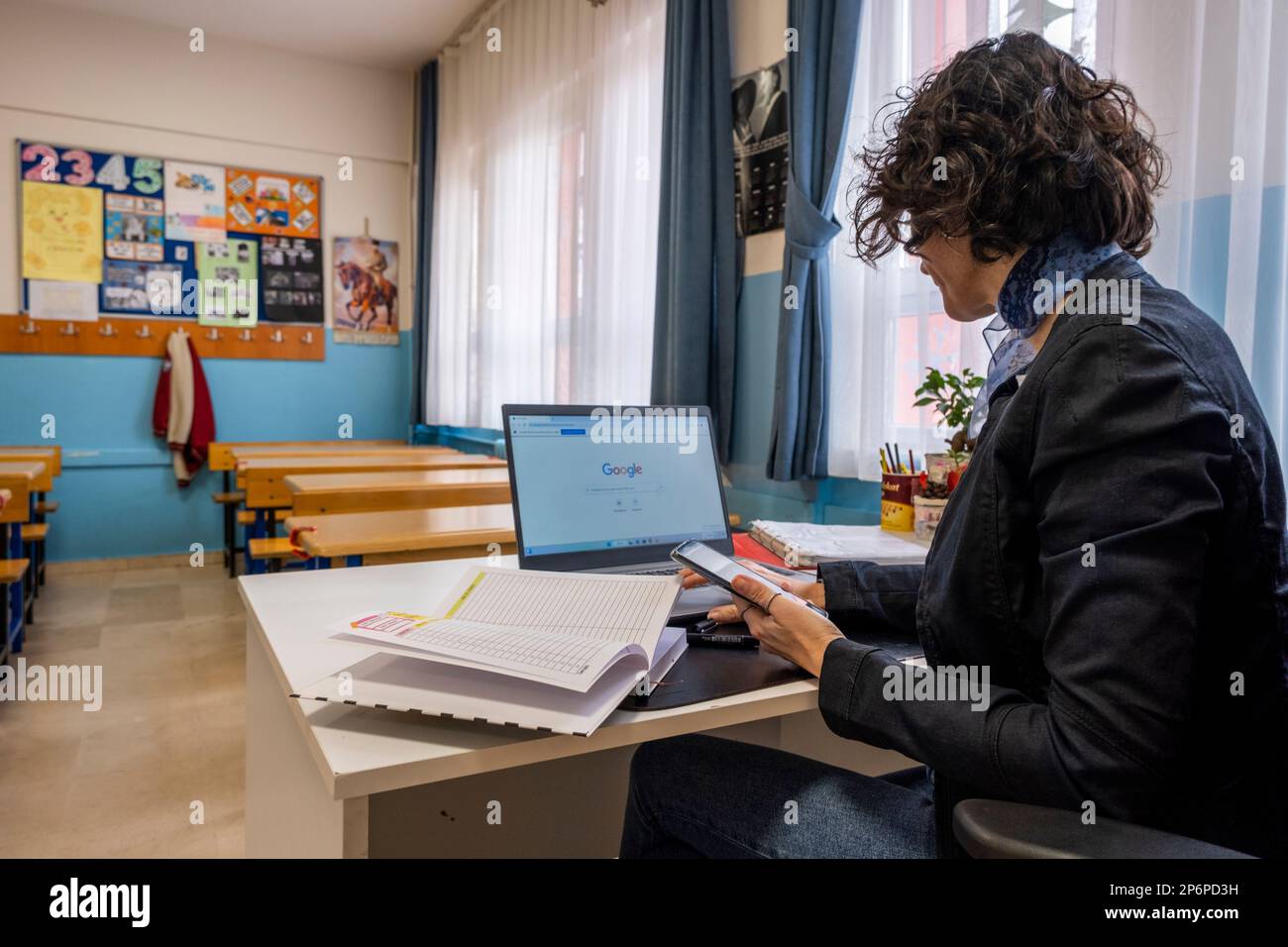 Die Rückansicht einer Lehrerin mit braunen kurzen Haaren und legerer Kleidung liest Notizen in ihrem Notizbuch in einem leeren Klassenzimmer. Horizontale Innenaufnahmen Stockfoto