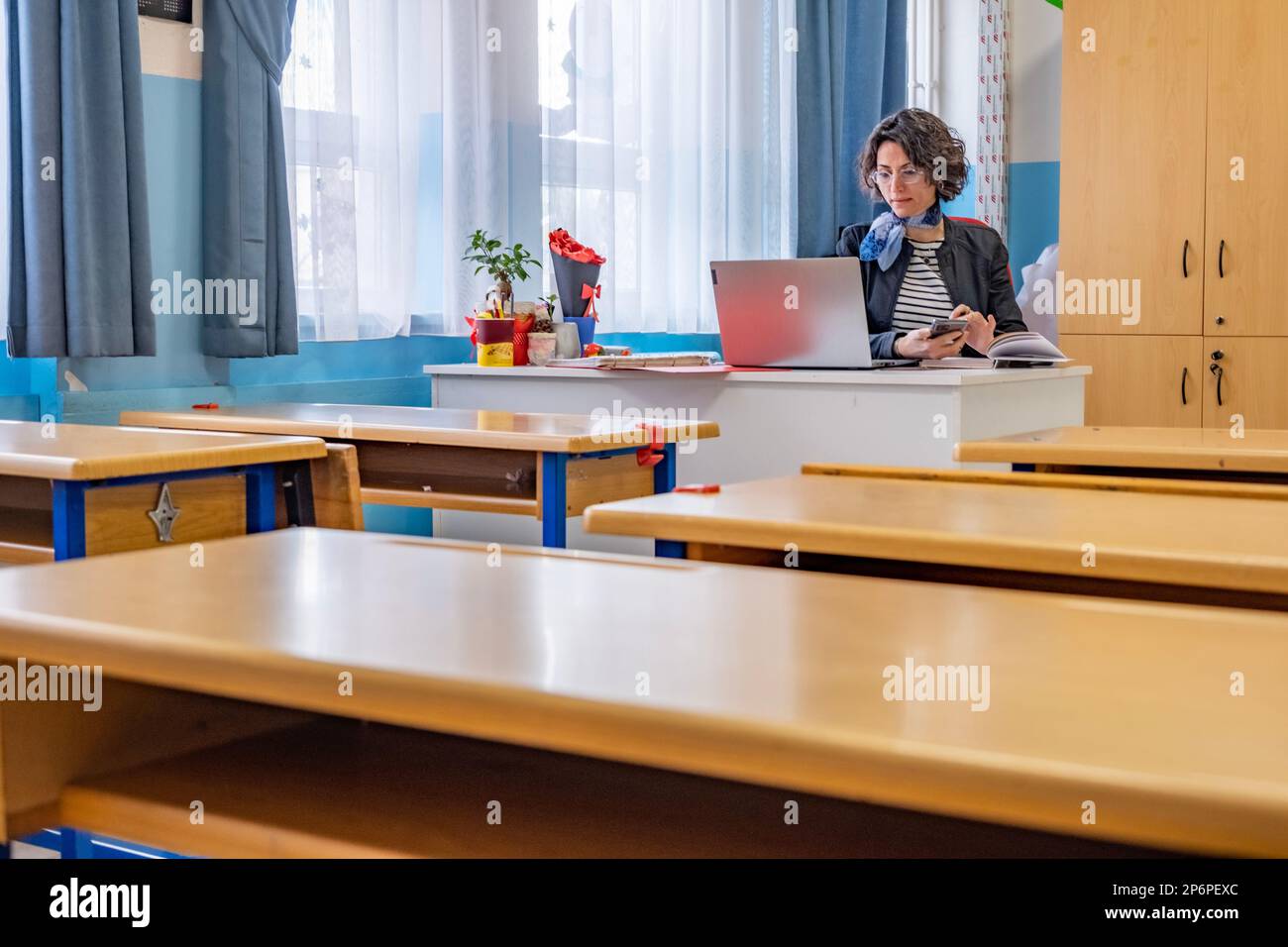 Reife Lehrerin schreibt Notizen in ihrem Notizbuch in leerem Klassenzimmer. Viele leere Tische vor ihr. Horizontale Innenaufnahmen mit Kopierbereich. Stockfoto