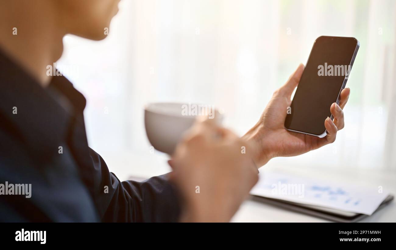 Zugeschnittenes und Nahaufnahme-Bild eines erfolgreichen und professionellen asiatischen Geschäftsmanns oder männlichen Chefs, der sein Telefon benutzt, während er an seinem Schreibtisch Kaffee trinkt i Stockfoto
