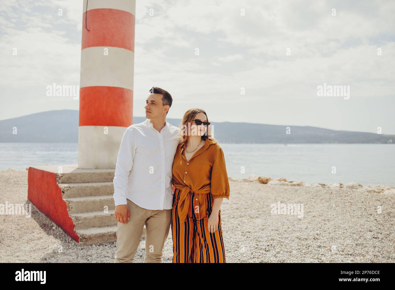 Ein Paar steht in der Nähe eines Leuchtturms am Strand vor dem Hintergrund des Meeres und der Berge. Urlaub, Flitterwochen, Reisen. Schickes Paar, das lacht Stockfoto