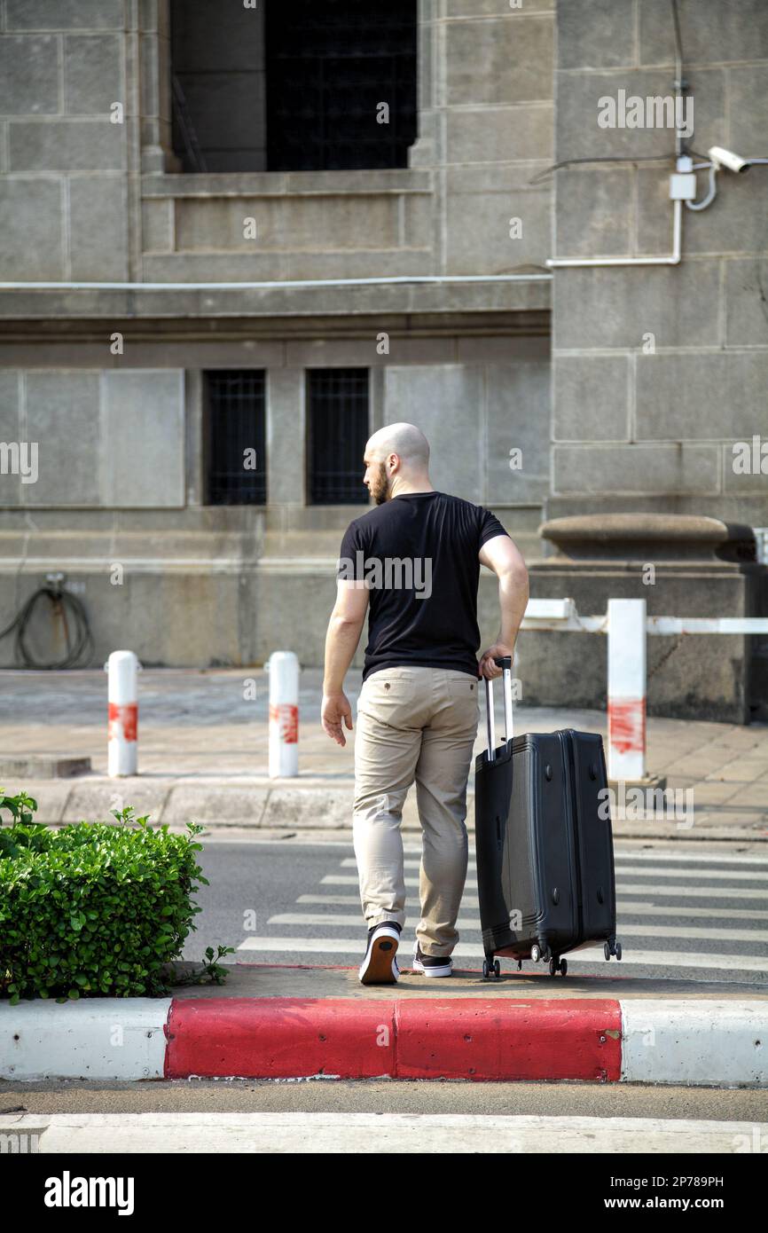 Ein junger Mann in legerer Kleidung überquert die Straße und hält einen Koffer von hinten gesehen. Glatzenkopf auf Geschäftsreise, der am Flughafen ankommt und zu Fuß unterwegs ist Stockfoto